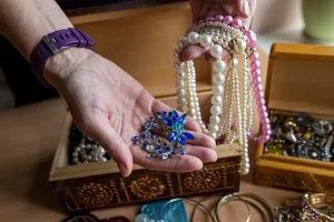 Hands holding a blue jeweled brooch and pearl necklaces from a wooden jewelry box at an estate sale by C'z Estate Sales LLC.